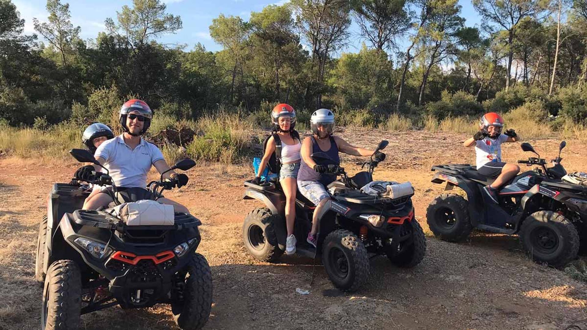Groupe de personnes en randonnée quad dans un paysage naturel, portant des casques de protection. Les quads sont noirs avec des touches de rouge, et l'arrière-plan est composé de végétation méditerranéenne sous un ciel dégagé.