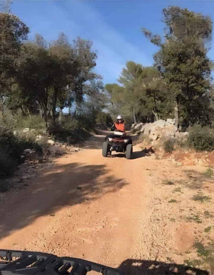 Randonnée en quad sur un sentier en pleine nature, entouré d’arbres et de végétation méditerranéenne sous un ciel bleu dégagé. Un pilote casqué roule sur un chemin de terre avec un autre quad visible au premier plan.