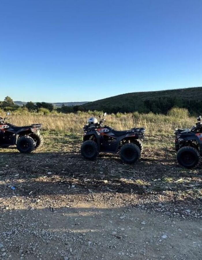 Randonnée en quad sur un sentier en pleine nature, entouré d’arbres et de végétation méditerranéenne sous un ciel bleu dégagé. Un pilote casqué roule sur un chemin de terre avec un autre quad visible au premier plan.