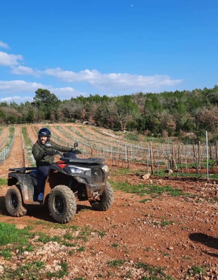 Randonnée en quad sur un sentier en pleine nature, entouré d’arbres et de végétation méditerranéenne sous un ciel bleu dégagé. Un pilote casqué roule sur un chemin de terre avec un autre quad visible au premier plan.